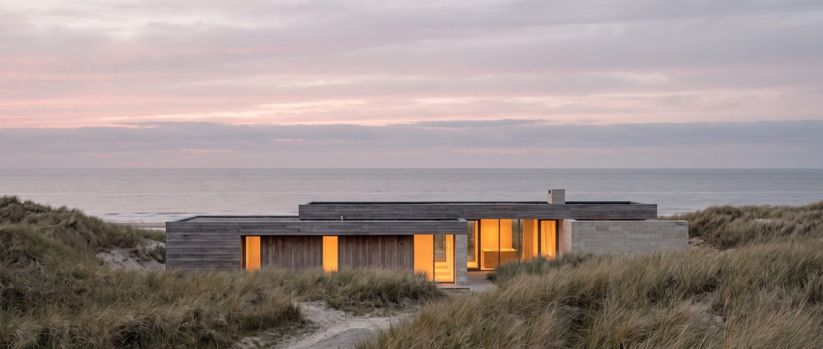 Duinstilte bij schemering: een lage villa van hout en kalksteen in de duinen, met warm verlichte ramen en de Noordzee daarachter.